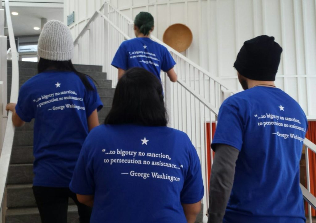 Four models wearing the "Refugees Welcome" t-shirt graphic, designed for the Worcester Refugee Assistance Project (Worcester, Massachussetts, USA). Desiger: Ben Young Landis/Creative Externalities. The photo shows the back of the t-shirt, which is a royal blue. The graphic reads: "to bigotry no sanction, to persecution no asssistance. George Washington" in white ink, with a white star graphic.