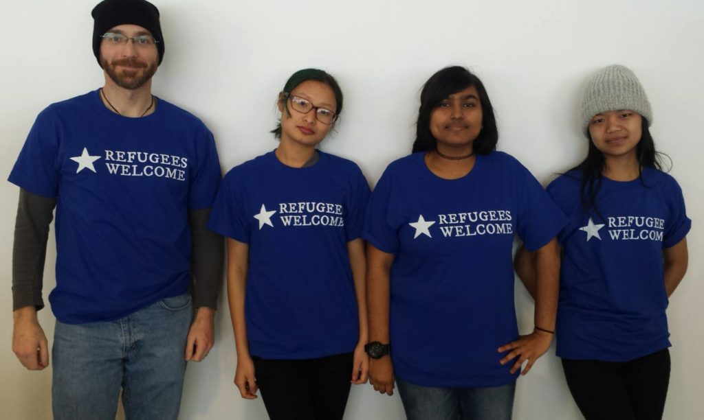 Four models wearing the "Refugees Welcome" t-shirt graphic, designed for the Worcester Refugee Assistance Project (Worcester, Massachussetts, USA). Desiger: Ben Young Landis/Creative Externalities. The photo shows the front of the t-shirt, which is a royal blue. The graphic reads: "Refugees Welcome" in white ink, with a white star graphic.