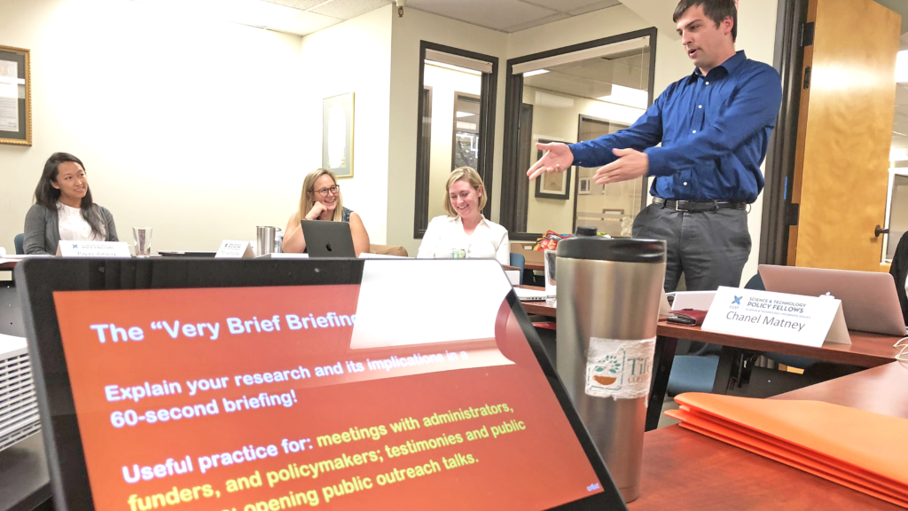 A scene from a "Connecting with Your Audience" Workshop taught by Ben Young Landis for the CCST Science & Technology Policy Fellowship. Here, 2019 CCST Science Fellow Jared Ferguson gestures during his practice briefing in front of colleagues. Jared is wearing a French blue dress shirt and gray trousers. A laptop screen shows the text "The Very Brief Briefing".