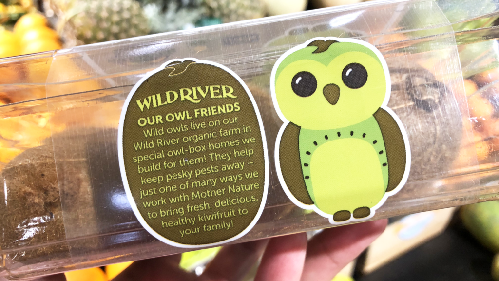 Our Owl Friends and associated kiwi owl logos are trademarks of Wild River Marketing Inc. Designed by Ben Young Landis and Guy Rogers. The photo shows the front side of a Wild River plastic clamshell container for kiwifruit. Next to a green kiwi owl, the text reads: Our Owl Friends: Wild owls live on our Wild River organic farm in special owl-box homes we build for them! They help keep pesky pests away — just one of many ways we work with Mother Nature to bring fresh, delicious kiwifruit to your family!