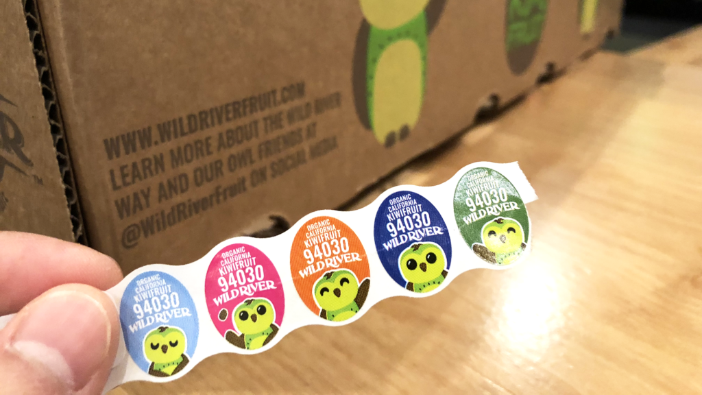 Our Owl Friends and associated kiwi owl logos are trademarks of Wild River Marketing Inc. Designed by Ben Young Landis and Guy Rogers. The photo shows a series of P L U stickers created for Wild River kiwifruit. Each oval sticker has a green kiwi owl in a pose (respectivul, playful, waving, standing, waving), against blue, pink, orange, dark blue, and green backgrounds. The stickers read: Organic California Kiwifruit 94030 Wild River.