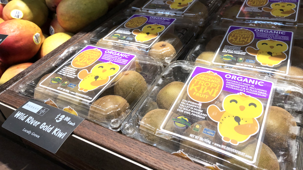 Our Owl Friends and associated kiwi owl logos are trademarks of Wild River Marketing Inc. Designed by Ben Young Landis and Guy Rogers. The photo shows plastic clamshell containers with gold kiwifruit, stocked on the shelf of Market 5 One 5 in Sacramento, California. The container's graphic shows the smiling Wild River Tropikiwi owl with one wing holding a spoon. Graphic says "California's Sacramento Valley is home to Wild River and Our Owl Friends!" and that the fruits are organic, naturally high in antioxidants, including vitamins C & E! Plus fiber, potassium & magnesium!